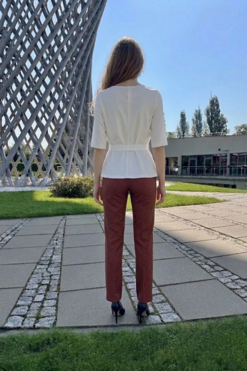 Na modelce stojącej tyłem biała bluzka z paskiem materiałowym, w tym samym kolorze co bluzka, z rękawami o długości 3/4, i niewielkim okrągłym dekoltem, na karku kryte pęknięcie zapinane na guzik