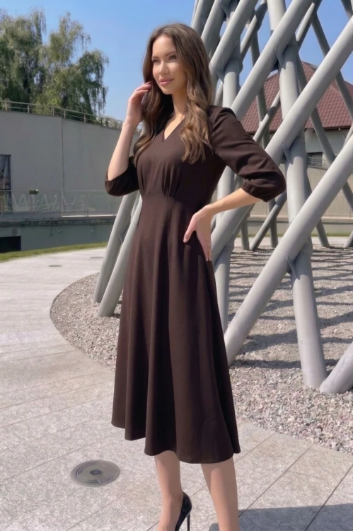 Na modelce stojącej przodem, sukienka w kolorze ciemnobrązowym, o długości za kolana, lekko rozkloszowana, z odcięciem pod biustem w kształcie trójkąta, z lekkim przymarszczeniem, dekolt w literkę v, rękaw o długości 3/4 zakończony gumką.