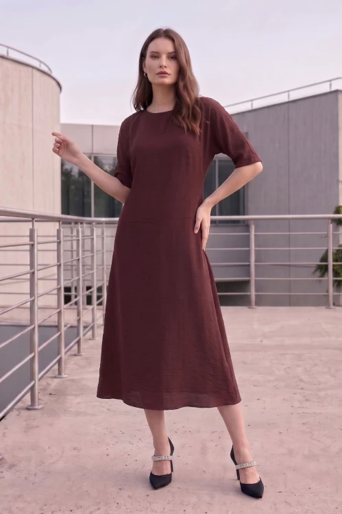Na modelce stojącej przodem, sukienka midi w kolorze brązowym, z obniżoną talią, niewielki okrągły dekolt, rękawek kimonowy o długości do łokcia, dół sukienki  rozkloszowany