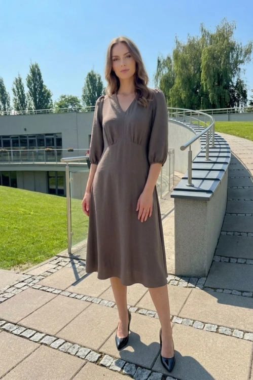 Na modelce, sukienka w kolorze fango, odcień brązu, o długości za kolana, lekko rozkloszowana, z odcięciem pod biustem w kształcie trójkąta, z lekkim przymarszczeniem, dekolt w literkę v, rękaw o długości 3/4 zakończony gumką.