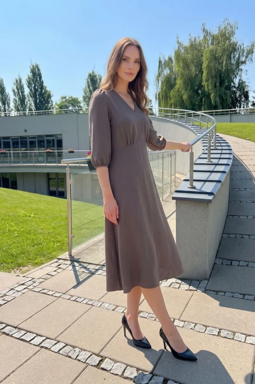 Na modelce, sukienka w kolorze fango, odcień brązu, o długości za kolana, lekko rozkloszowana, z odcięciem pod biustem w kształcie trójkąta, z lekkim przymarszczeniem, dekolt w literkę v, rękaw o długości 3/4 zakończony gumką.