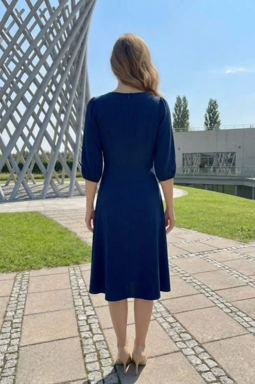 Na modelce stojącej tyłem, granatowa sukienka, o długości za kolana, lekko rozkloszowana, rękaw o długości 3/4 zakończony gumką, zapinana na kryty suwak