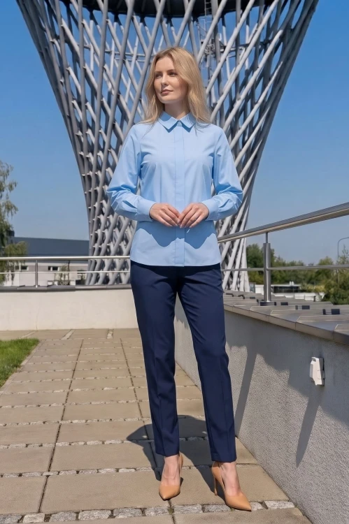Na modelce błękitna koszula damska, o prostym kroju, z klasycznym kołnierzykiem, zapinana na guziki ukryte pod pionową listwą. Koszula ma długie rękawy zakończone mankietami zapinanymi na dwa guziki.