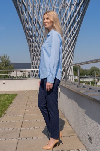 Na modelce stojącej bokiem błękitna koszula damska, o prostym kroju, z klasycznym kołnierzykiem, zapinana na guziki ukryte pod pionową listwą. Koszula ma długie rękawy zakończone mankietami zapinanymi na dwa guziki.
