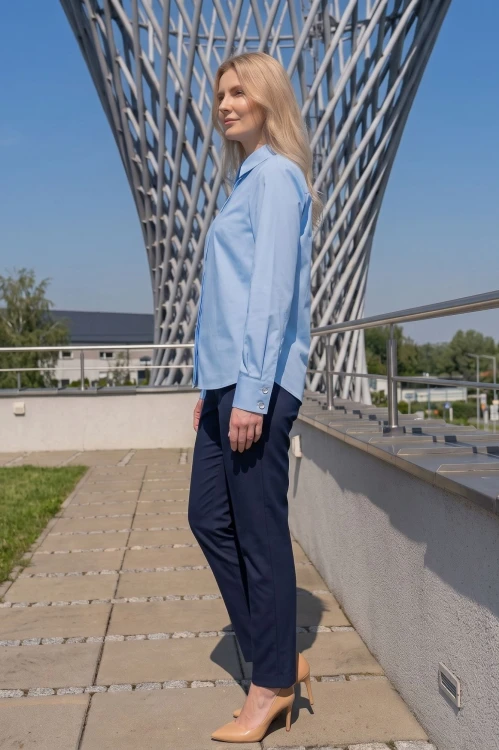 Na modelce stojącej bokiem błękitna koszula damska, o prostym kroju, z klasycznym kołnierzykiem, zapinana na guziki ukryte pod pionową listwą. Koszula ma długie rękawy zakończone mankietami zapinanymi na dwa guziki.