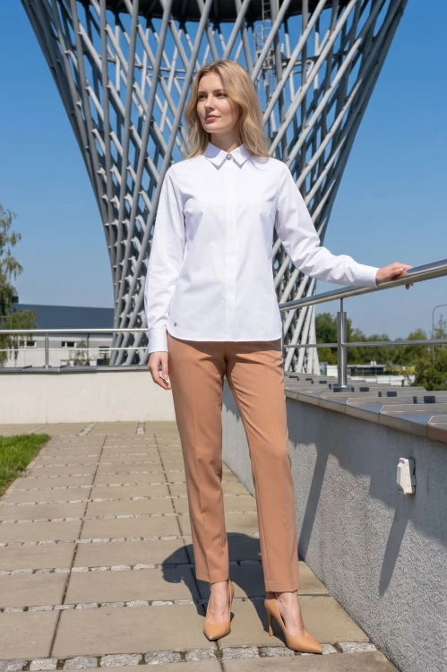 Na modelce elegancka biała koszula damska, o prostym kroju, z klasycznym kołnierzykiem, zapinana na guziki ukryte pod pionową listwą. Koszula ma długie rękawy zakończone mankietami zapinanymi na dwa guziki.