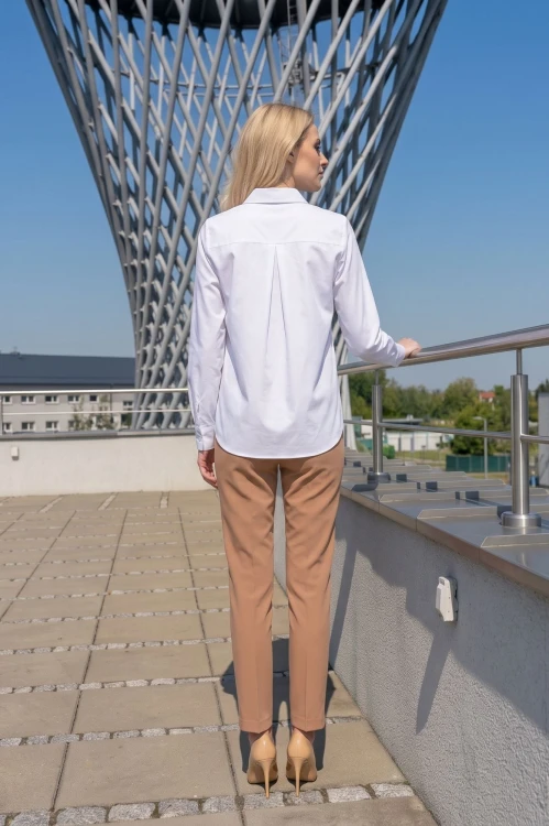 Na modelce stojącej tyłem elegancka biała  koszula damska, o prostym kroju, z klasycznym kołnierzykiem. Ma długie rękawy zakończone mankietami zapinanymi na dwa guziki. Na plecach karczek, poniżej którego znajduje się kontrafałda