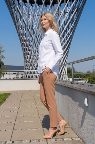 Na modelce elegancka biała koszula damska, o prostym kroju, z klasycznym kołnierzykiem, zapinana na guziki ukryte pod pionową listwą. Koszula ma długie rękawy zakończone mankietami zapinanymi na dwa guziki.