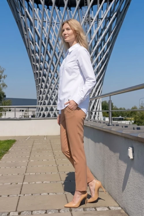 Na modelce elegancka biała koszula damska, o prostym kroju, z klasycznym kołnierzykiem, zapinana na guziki ukryte pod pionową listwą. Koszula ma długie rękawy zakończone mankietami zapinanymi na dwa guziki.