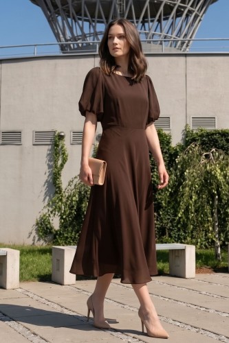 Na modelce sukienka o długości midi  rozkloszowana w kolorze brązowym, z okrągły dekoltem, odcięciem po wyżej talii, krótkim rękawkiem zakończonym gumką, tworząc bufkę.