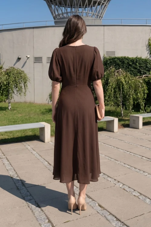 Na modelce stojącej tyłem sukienka o długości midi  rozkloszowana w kolorze brązowym z odcięciem po wyżej talii, krótkim rękawkiem zakończonym gumką, tworząc bufkę, zapinana na kryty suwak