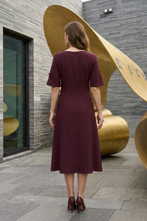 Na modelce stojącej tyłem, sukienka midi w kolorze burgundowym, z obniżoną talią, rękawek kimonowy o długości do łokcia,  lekko rozkloszowany, zapinana na kryty suwak